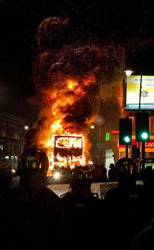 A double decker bus burns as riot police look on